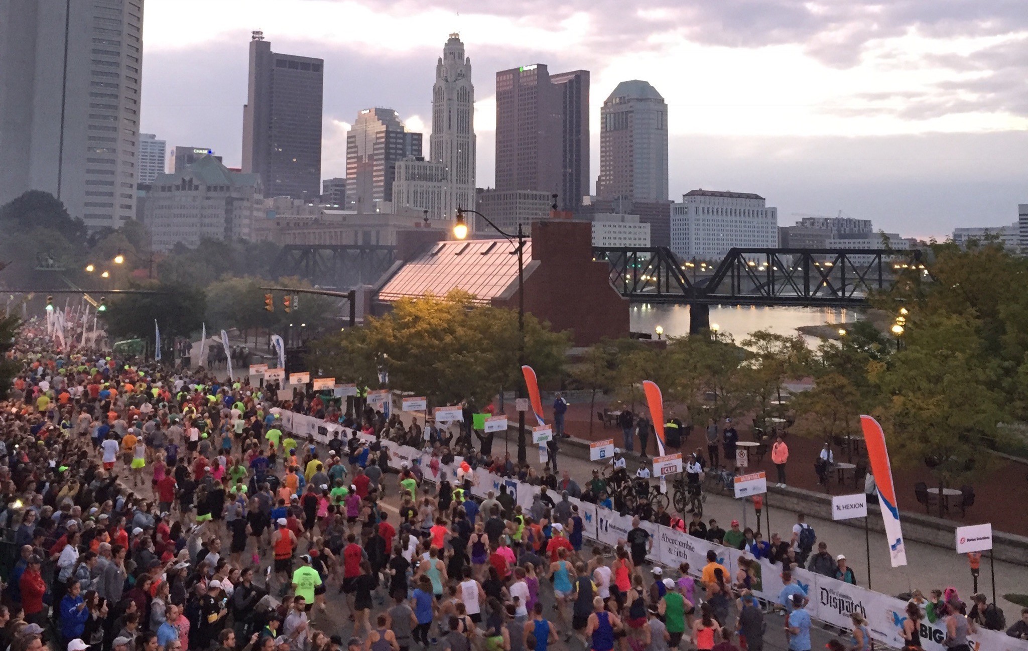 Thousands of marathon runners on a city street beside North Bank Park Pavilion with the Columbus skyline and Scioto River in the background.
