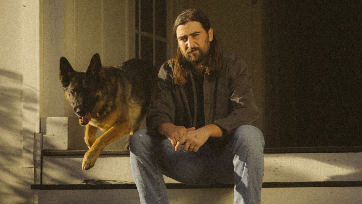 Noah Kahan with beard and shoulder-length hair sitting on porch steps next to a German Shepherd dog in warm sunlight.