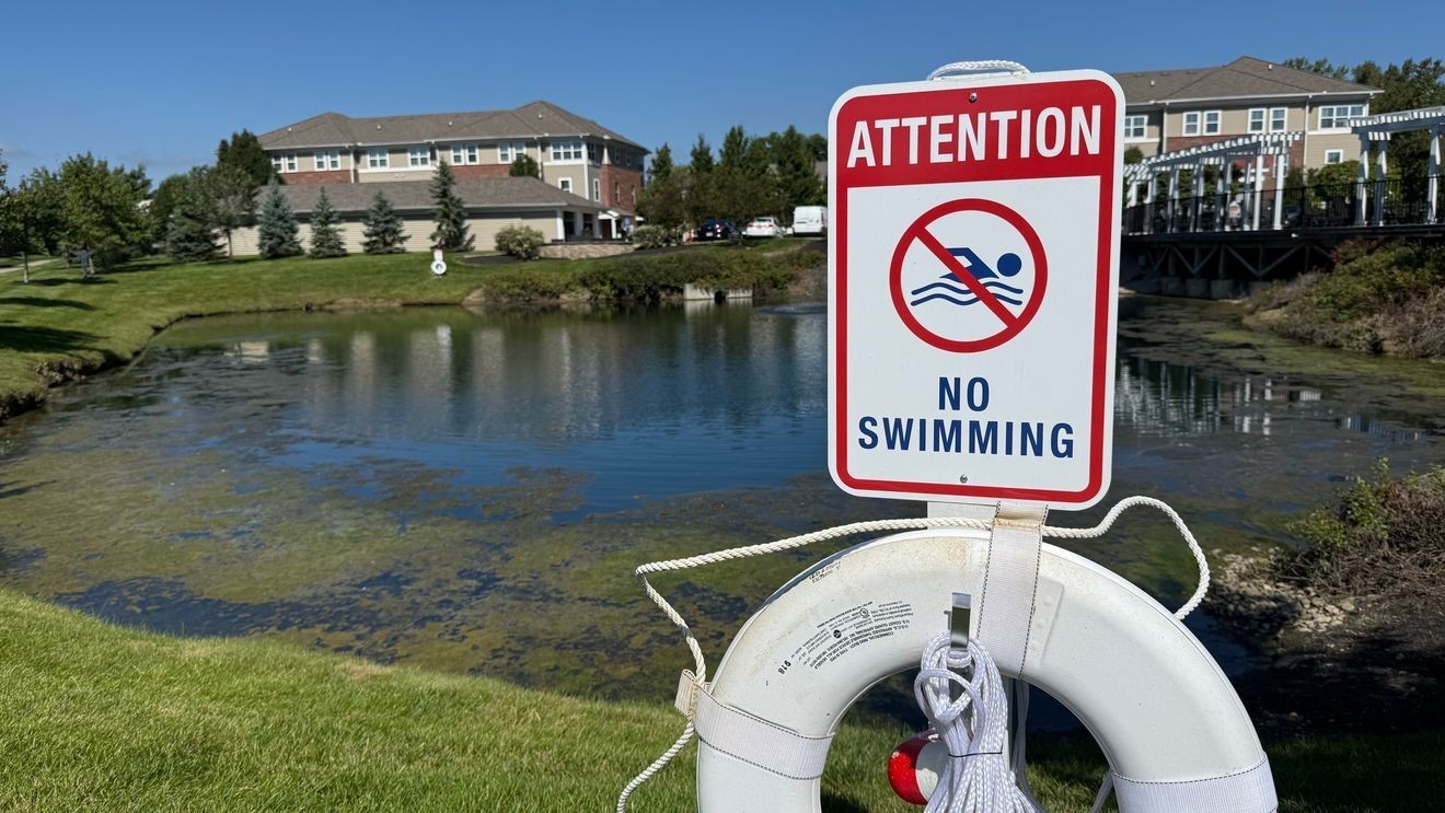 A white life buoy and red-and-white Attention No Swimming warning sign posted along the grass bank of a residential retention pond in Columbus, with apartment buildings in the background.