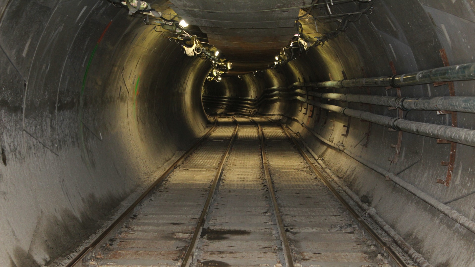 Interior view of a circular precast concrete deep tunnel with narrow-gauge tracks running down the center, illuminated by overhead lights.