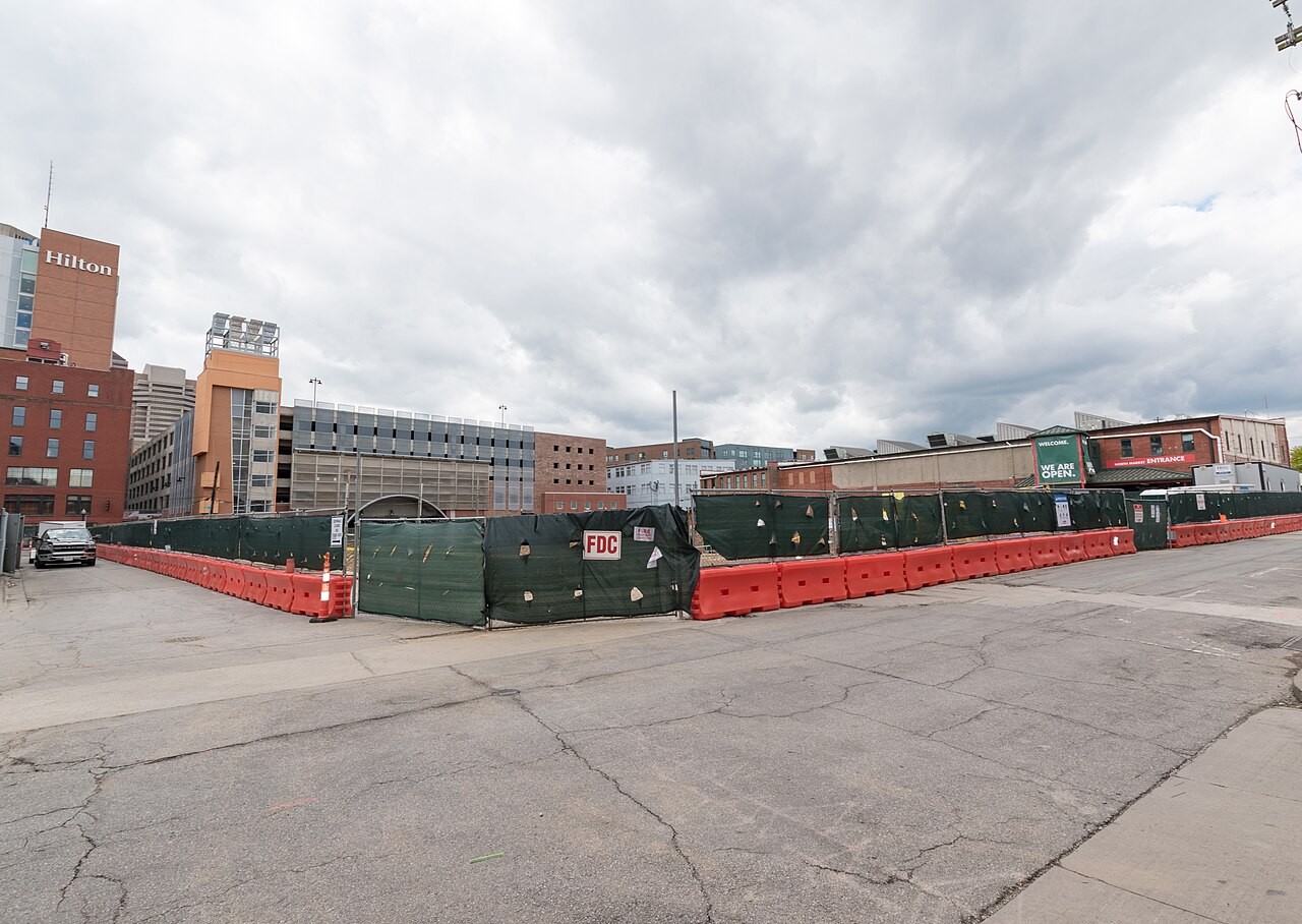Exterior ground-level view of the Merchant Building construction site enclosed by orange barriers and green fencing, adjacent to the North Market building in Columbus.