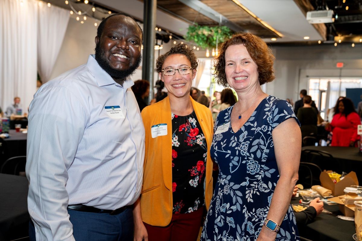 Three attendees smiling together at a Columbus Metropolitan Club forum, with event tables and string lights in the background.