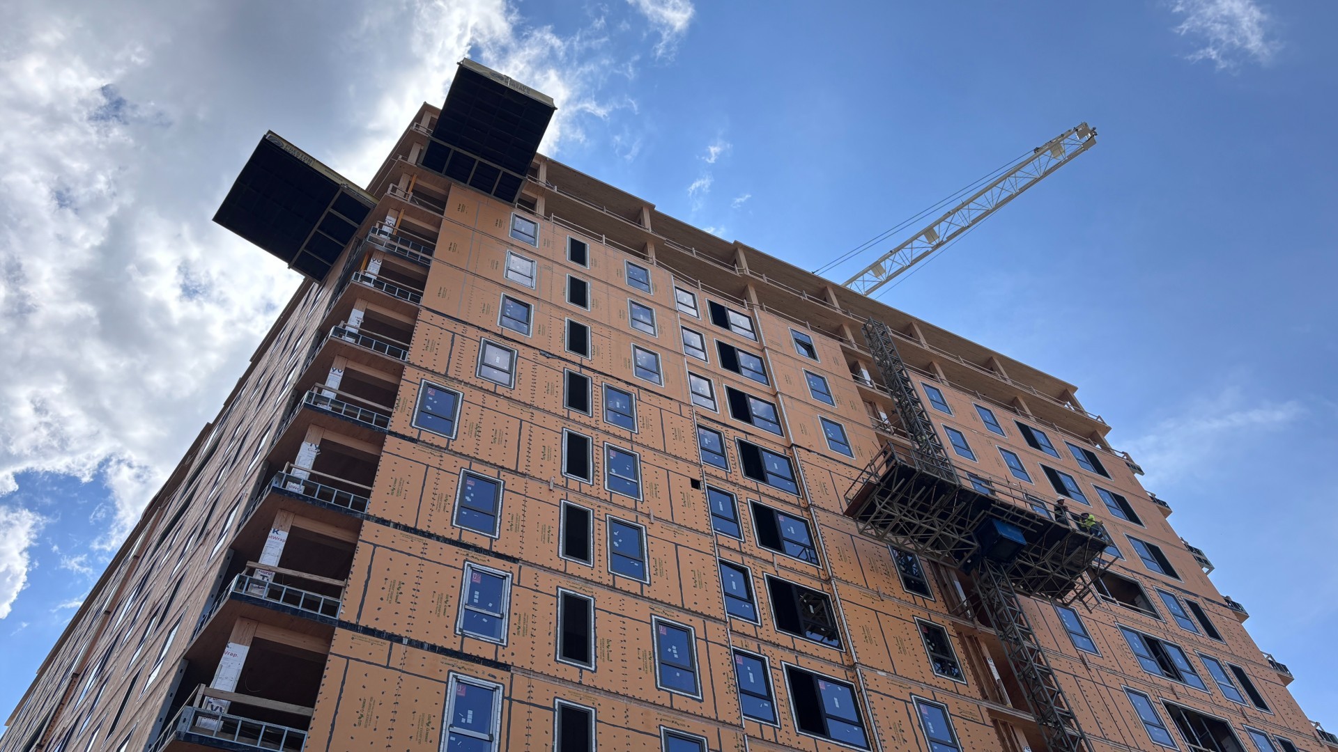 Low-angle view of the 13-story INTRO Columbus mass timber student tower under construction, showing orange weather-barrier sheathing, exterior hoist, and a crane boom.