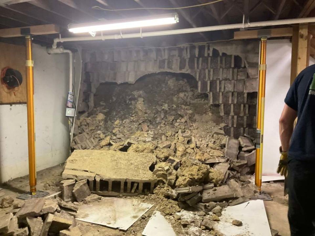A collapsed basement wall of concrete blocks and earth inside 2 South Vernon Street, with yellow steel shoring jacks supporting ceiling beams.