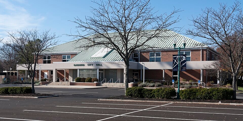 Exterior view of Gahanna City Hall, a red brick municipal building with a green metal roof fronted by a paved parking lot.