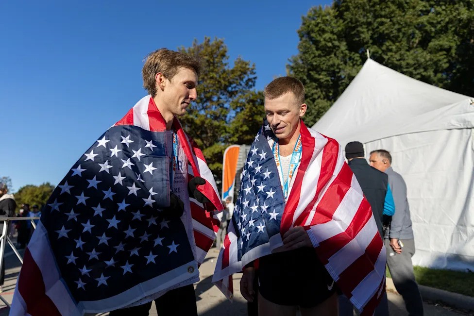 Two male marathon finishers draped in American flags and wearing medals converse outside post-race tents under clear blue skies.