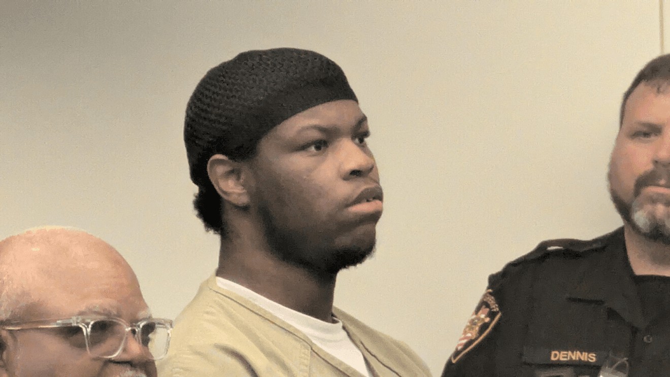Tamari Stroughter in a black skullcap and tan correctional jumpsuit standing in court with a sheriff's deputy.