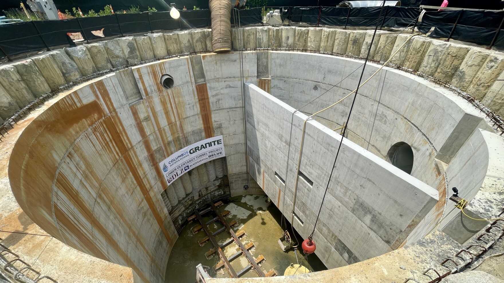 High-angle wide shot looking down into a deep concrete-lined drop shaft with internal dividing walls, tunnel portal openings, and project signage.