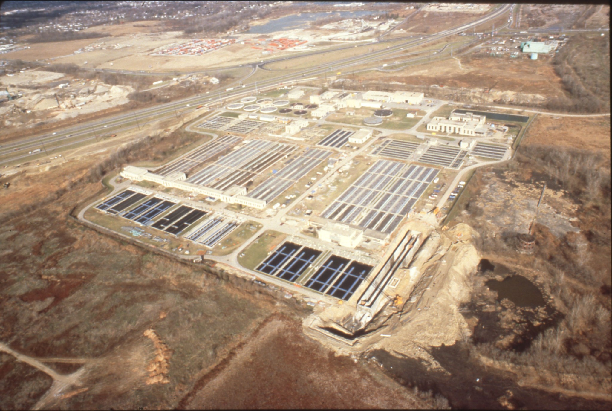 High-angle aerial photograph of the Jackson Pike Wastewater Treatment Plant in Columbus, Ohio, showing large rectangular aeration and settling basins, circular digester tanks, administrative buildings, and Interstate 71 in the background.