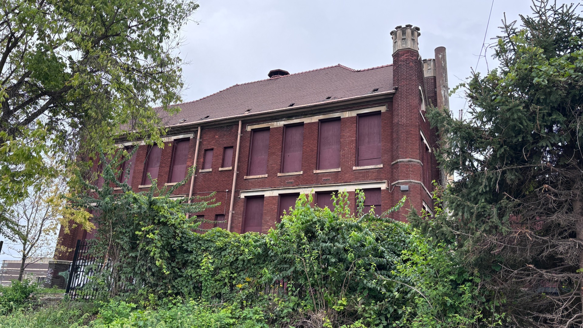 Exterior facade of the vacant Holy Family School building in Franklinton, featuring dark red brick, boarded windows, decorative corner chimneys, and dense foreground greenery.