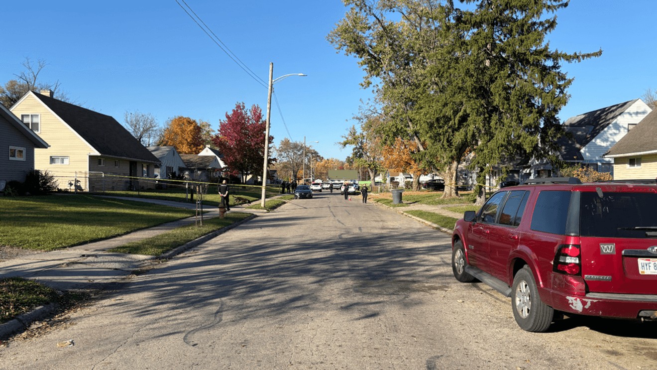 Columbus police officers and yellow crime scene tape cordoning off a residential street on Marcia Drive with parked vehicles and homes in the background.
