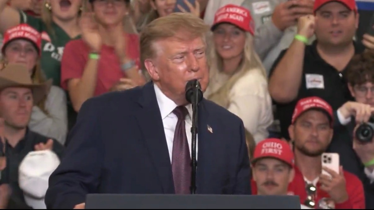 Donald Trump speaks at a podium microphone during a midterm campaign rally with supporters wearing red hats behind him.