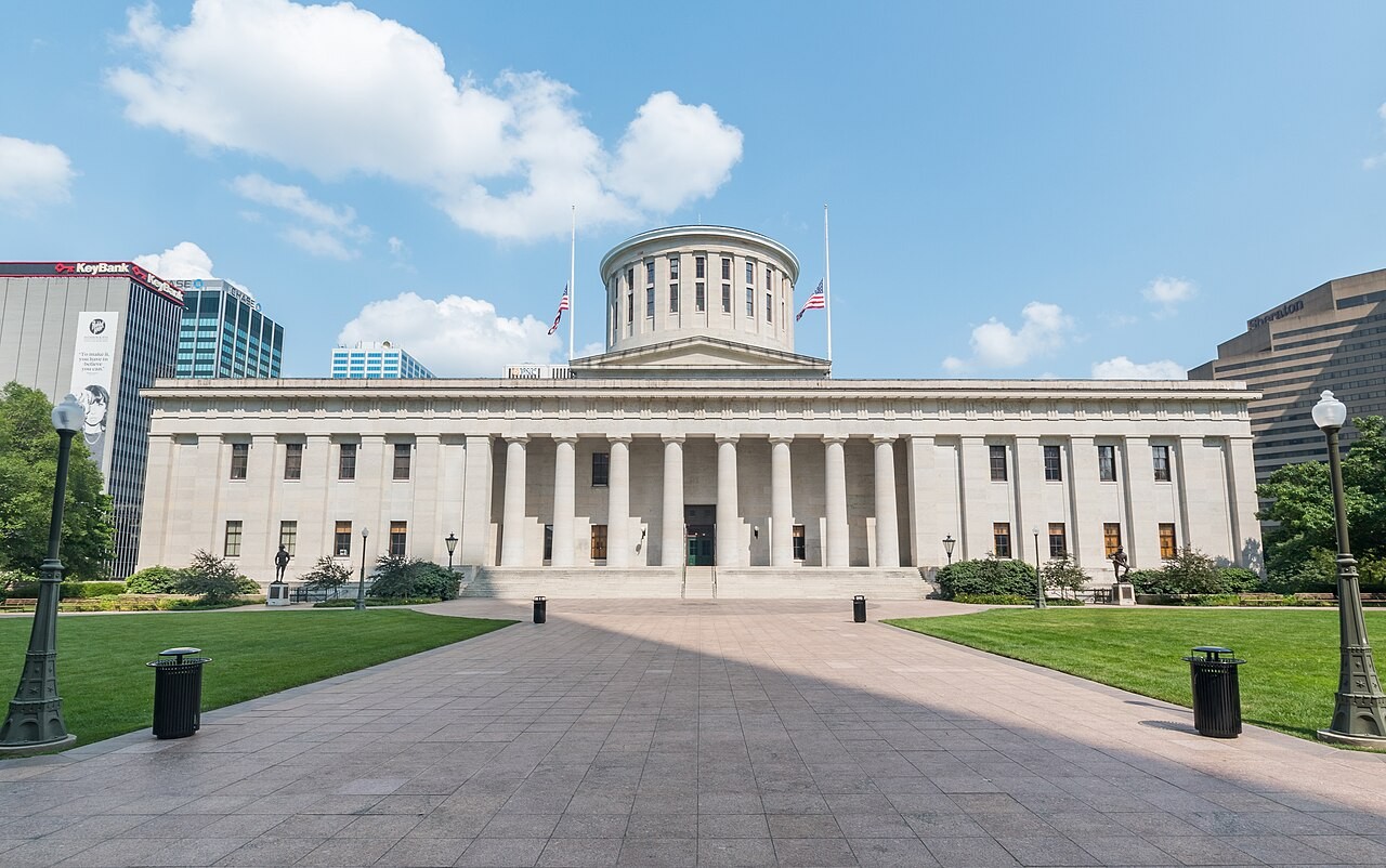 Frontal daylight view of the Greek Revival Ohio Statehouse facade and rotunda drum across the empty paved plaza of Capitol Square in Columbus.