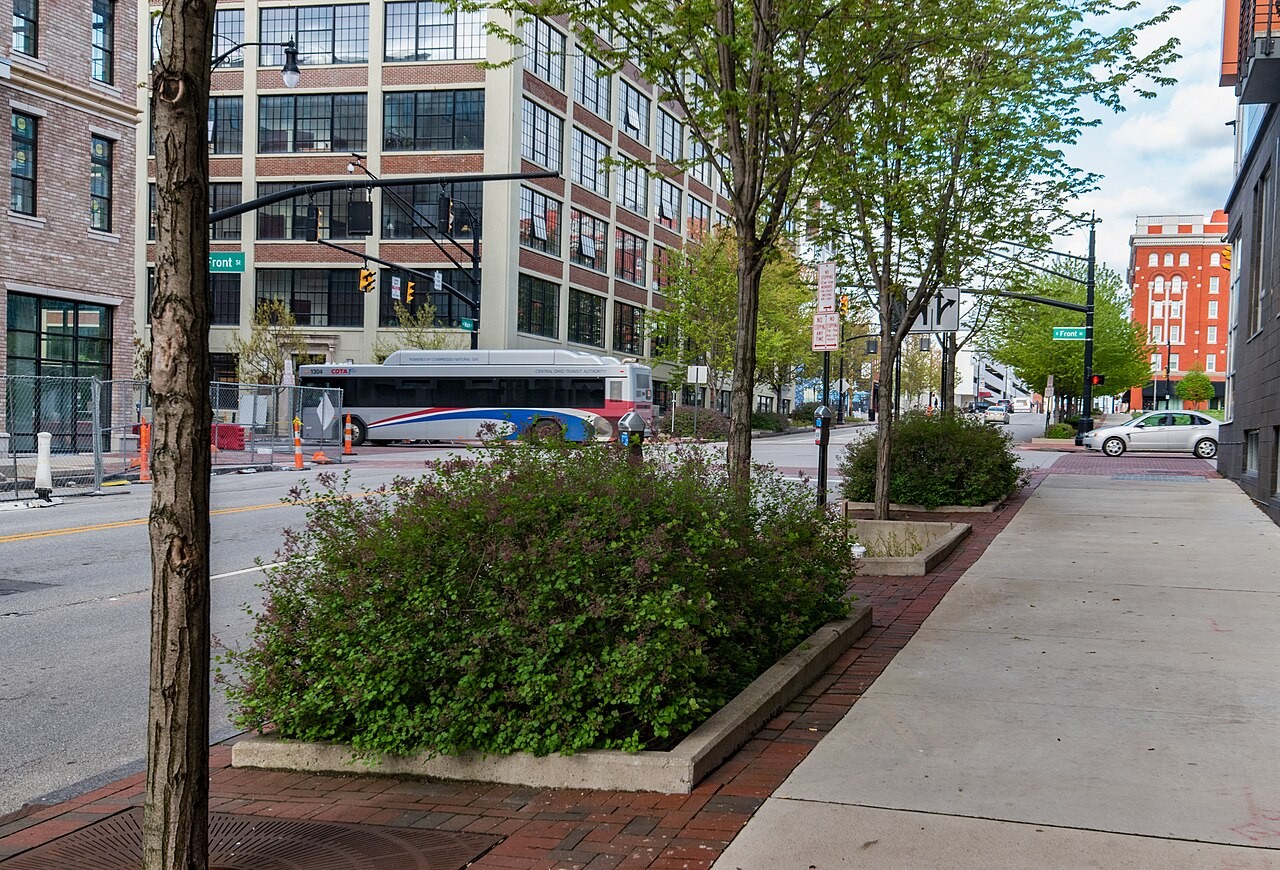 Planted curbside bioretention basin with dense green shrubs and young trees along a Columbus sidewalk, with a COTA transit bus and multistory buildings in the background.