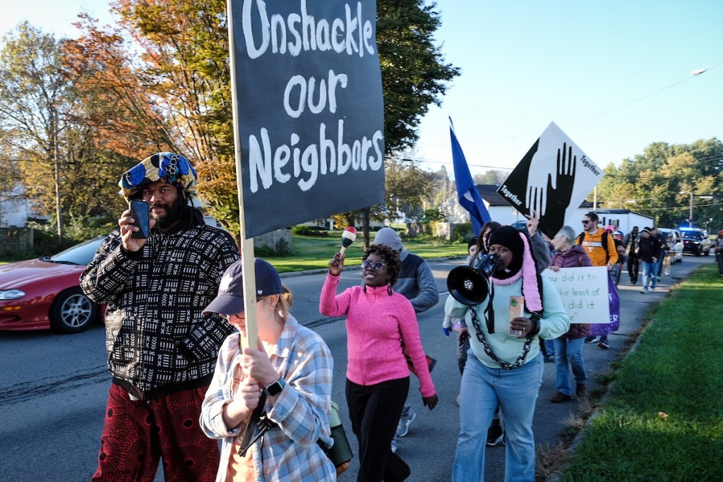 Advocates and community members march down an Ohio street carrying a large black sign reading 'Unshackle our Neighbors', accompanied by a woman with a megaphone and percussion instrument.