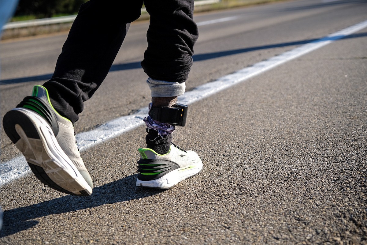Close-up shot of a person's lower legs and running shoes walking on a paved road shoulder, with a black electronic GPS monitoring device strapped around the right ankle.
