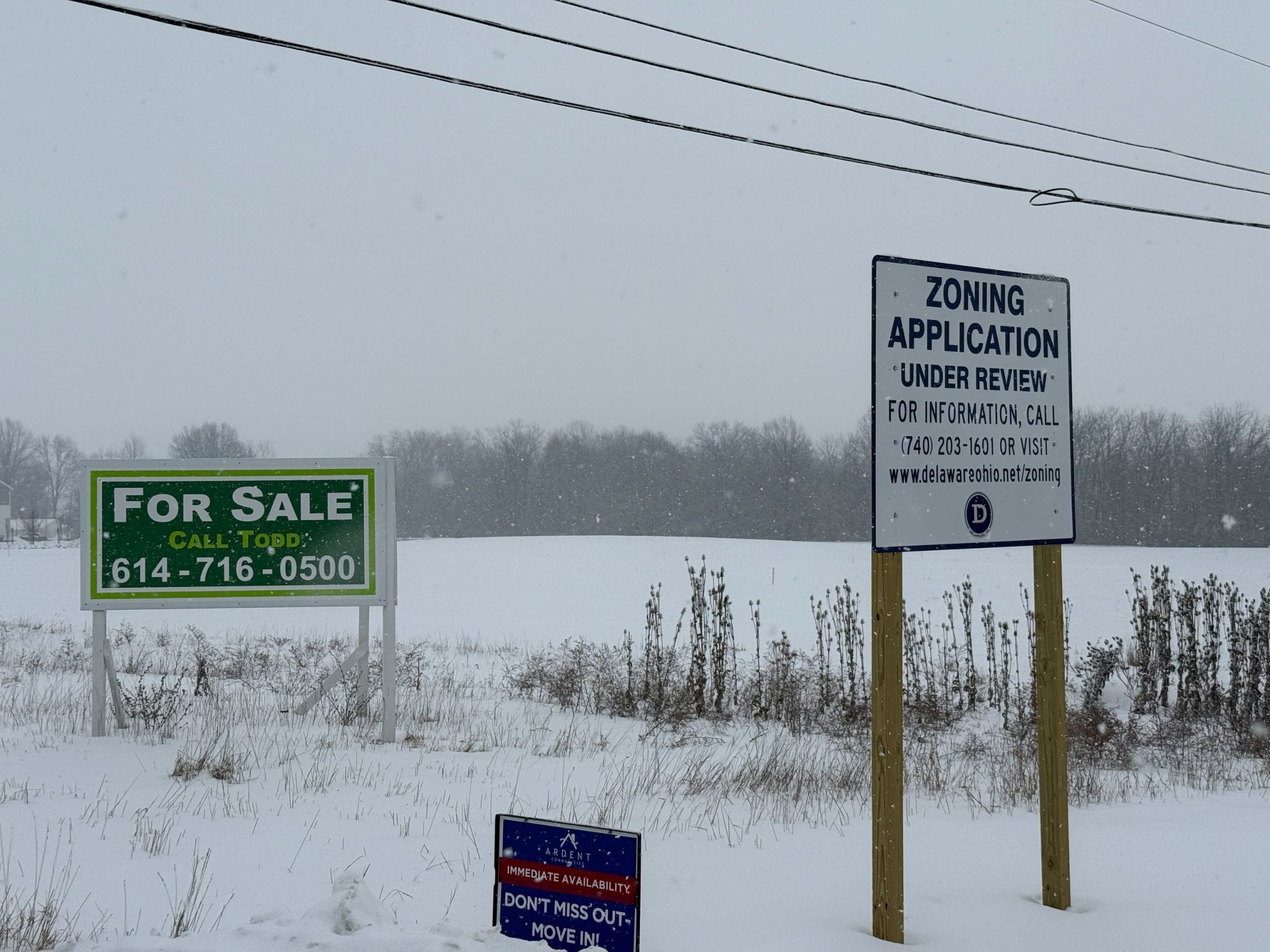 A snow-covered field along Sunbury Road in Delaware, Ohio, featuring a City of Delaware zoning review sign and a real estate sign.