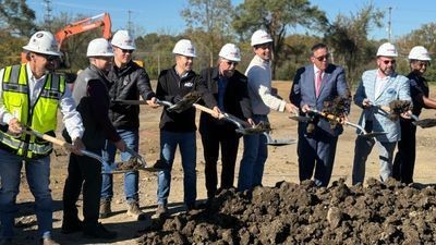 City officials and police leadership wearing white hard hats hold shovels in a dirt mound during a groundbreaking ceremony.