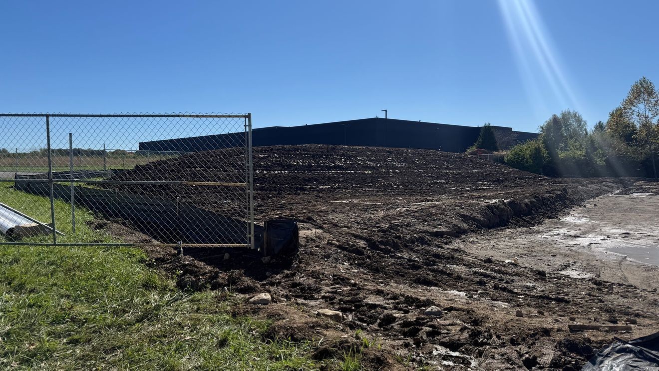 A graded dirt construction site with a large mound of soil behind a chain-link fence under a clear blue sky.