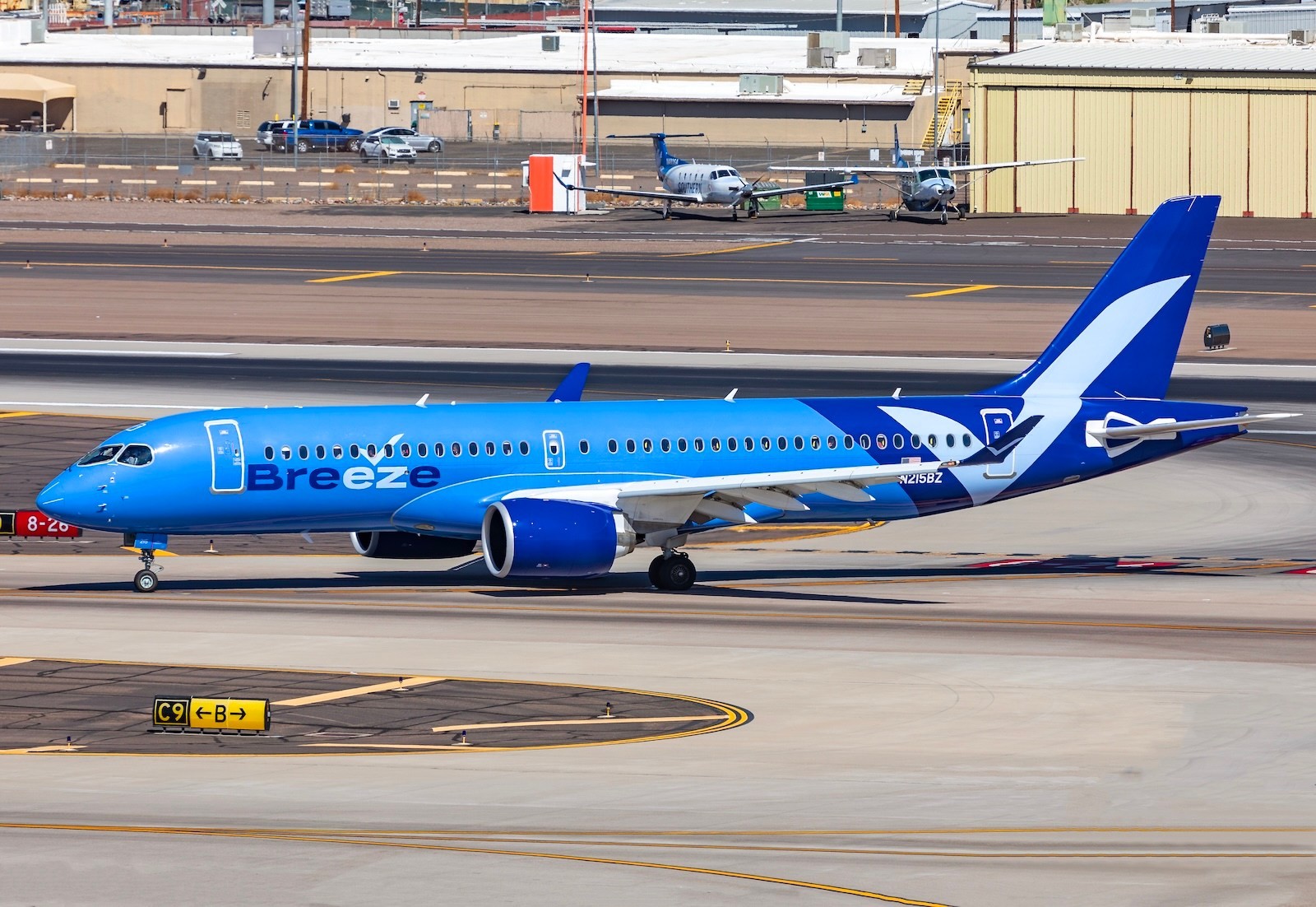 A Breeze Airways Airbus A220 passenger aircraft in blue livery taxiing along an airport taxiway with runway markings and distant hangars.